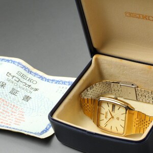 May include: A gold Seiko watch with a rectangular face and a gold strap sits in a blue box with a beige interior. The watch is displayed with a matching gold strap. A warranty card with Japanese text is visible.