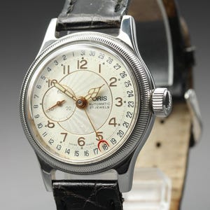 Puede incluir: Un reloj de pulsera automático Oris de acero inoxidable con esfera color crema, patrón guilloché, números arábigos, aguja de fecha y subesfera. El reloj presenta un bisel estriado y una correa de cuero negro con acabado texturizado.