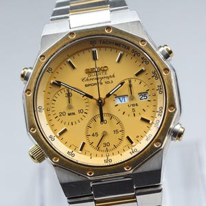 May include: A Seiko Quartz Chronograph Sports 100 watch with a gold-toned dial and a two-tone silver and gold stainless steel link bracelet. The watch features a tachymeter bezel, three sub-dials, a date window, and gold-toned hands and markers.