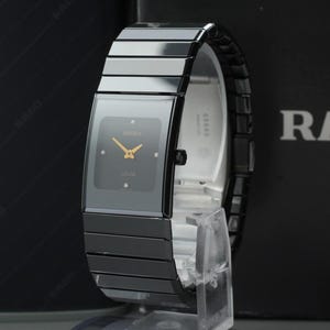 Könnte beinhalten: Eine Rado Armbanduhr mit einem rechteckigen schwarzen Keramikgehäuse und einem passenden schwarzen Keramikgliederarmband. Das schwarze Zifferblatt zeigt goldfarbene Zeiger und vier kleine goldfarbene Stundenmarkierungen. Das Rado-Logo ist sichtbar.