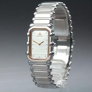 May include: A silver-toned Credor wristwatch with a rectangular face and a matching bracelet. The watch face is white with gold-colored hands and markers. The watch band is made of rectangular links.
