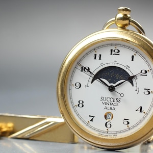 May include: A gold-coloured vintage pocket watch with a white face, black hands, and black numerals. The watch face displays the text "SUCCESS VINTAGE ALBA". A gold chain and tie clip are visible.