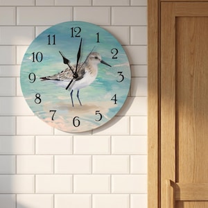 May include: A round wall clock featuring a painting of a sandpiper bird standing on a sandy beach with light blue water. The clock face has black Arabic numerals from one to twelve and black hour, minute, and second hands centred on the bird.