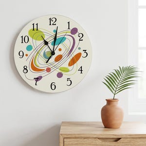May include: A round wall clock with an off-white face featuring black numerals from 1 to 12. The design includes black concentric oval lines and scattered abstract shapes in lime green, orange, purple, and teal. It has black hour and minute hands.