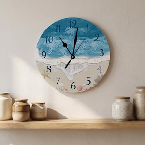 May include: A round wall clock featuring a printed ocean scene with blue waves crashing onto a sandy beach. The clock face displays dark blue numbers from 1 to 12, black hour and minute hands, and decorative illustrations of a starfish and seashells.
