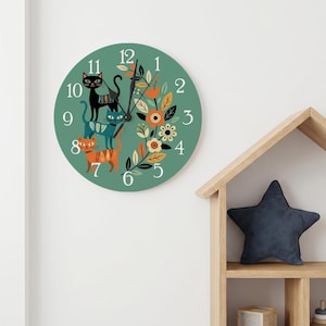 May include: A round wall clock with a sage green face featuring white numbers one through twelve. The design includes three stacked cats in black, teal, and orange with patterned bodies, alongside a floral wreath of orange and white flowers.