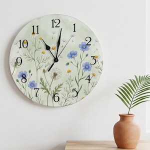 May include: A round wall clock with a pale green background featuring a botanical illustration of white daisies, blue flax flowers, and dandelion seed heads. The clock face has black Arabic numerals and black hour, minute, and second hands.