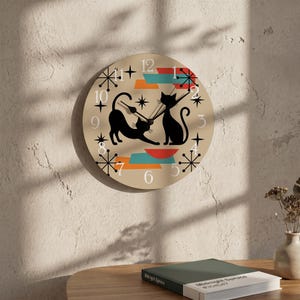 May include: A round wall clock with a beige face featuring two black cat silhouettes, one stretching and one sitting. The design includes white numbers, black starburst accents, and geometric shapes in orange and teal with black clock hands.