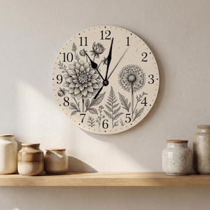 May include: A round wall clock with a cream-coloured face featuring black line art illustrations of a large dahlia, a dandelion, and fern leaves. The clock has black numerals from 1 to 12 and black hour, minute, and second hands.