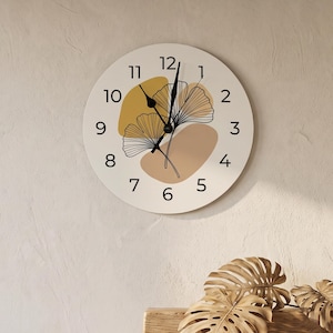 May include: A round wall clock with a cream-coloured face featuring black numerals from 1 to 12. The centre displays two black line-art ginkgo leaves layered over abstract mustard yellow and tan shapes. It has two black hands for timekeeping.