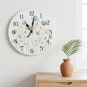 May include: A round wall clock with a white face featuring black numerals from 1 to 12. The clock face is decorated with a delicate illustration of light blue and sage green wildflowers on thin stems, accented by small gold dots and black specks.