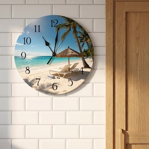 May include: A round wall clock featuring a tropical beach scene with white sand, turquoise water, palm trees, and a thatched umbrella over two lounge chairs. The clock face displays black Arabic numerals from 1 to 12 and black hour and minute hands.