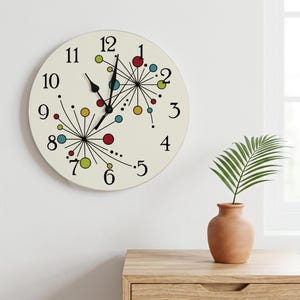 May include: A round wall clock with an off-white face featuring black numerals from 1 to 12. The design includes two black starburst patterns with small red, blue, yellow, and green circles at the ends of the lines. It has black hour and minute hands.