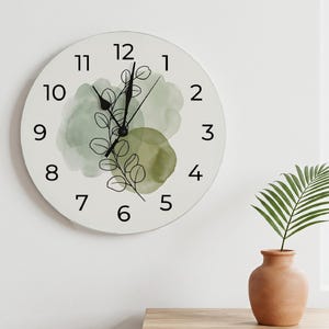Pode incluir: Um relógio de parede redondo com mostrador branco e algarismos arábicos pretos de 1 a 12. O centro apresenta um desenho de linha preta de um ramo com folhas sobreposto a formas abstratas de aguarela suave em tons de verde sálvia e oliva.