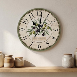 May include: A round wall clock with a sage green frame and a cream-colored face featuring black Arabic numerals. The center displays a botanical illustration of green and black olives on branches with the text Olives de Provence.