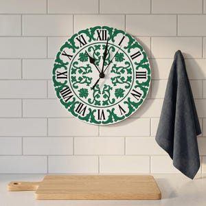 May include: A round wall clock featuring a white face with a green floral scroll pattern and black Roman numerals. The clock has two black hands and is positioned above a light-colored wooden cutting board and a dark gray hanging cloth.