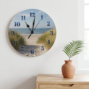 May include: A round wall clock featuring a scenic beach landscape with sand dunes and the ocean. The clock face displays dark blue numbers from 1 to 12 arranged in a circle, with black hour, minute, and second hands centred over the image.