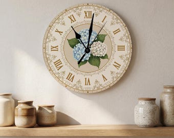 May include: A round wall clock with a beige face featuring Roman numerals and a central illustration of blue and white hydrangea flowers with green leaves. The clock has black hands and a decorative gold-toned filigree border pattern.