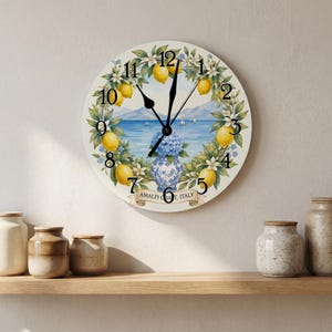 May include: A round wall clock featuring a coastal scene with a blue sea, mountains, and sailboats. A wreath of yellow lemons, green leaves, and white flowers surrounds a blue and white vase of blue hydrangeas. Text reads AMALFI COAST, ITALY.