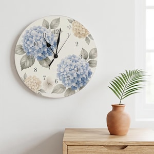 May include: A round wall clock with a cream-colored face featuring illustrations of blue and beige hydrangea flowers with green leaves. The clock has black numerals from 1 to 12 and thin black hour, minute, and second hands centered on the dial.