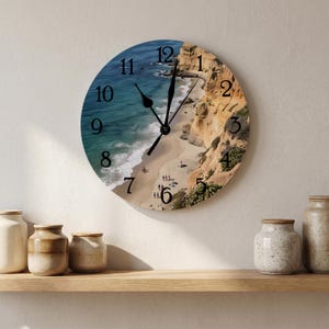 May include: A round wall clock featuring a photographic print of a sandy beach bordered by tall, tan-colored cliffs and blue ocean water. The clock face displays black Arabic numerals from one to twelve and two black hands indicating the time.