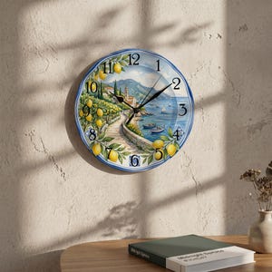 Könnte beinhalten: Eine runde Wanduhr mit einer Küstenszene, gelben Zitronen an Zweigen, einem Steinweg, blauem Meer, Booten und Bergen. Das Zifferblatt zeigt schwarze arabische Ziffern von 1 bis 12, schwarze Zeiger für Stunden und Minuten sowie einen blauen Schmuckrand.