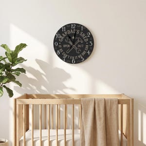 Puede incluir: Un reloj de pared redondo de color negro con números blancos y el texto "GRANDAD'S SHED" junto con "OPEN 24/7" y "IF HE CAN'T FIX IT NO ONE CAN". El centro muestra un gráfico de herramientas cruzadas, incluyendo una llave inglesa y un martillo, con manecillas negras.