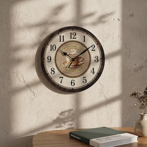 May include: A round wall clock with a dark brown frame and a cream-colored face featuring black Arabic numerals. The center displays a graphic of a coffee cup on a saucer with the text "WELCOME TO THE COZY CAFE" printed in black lettering.