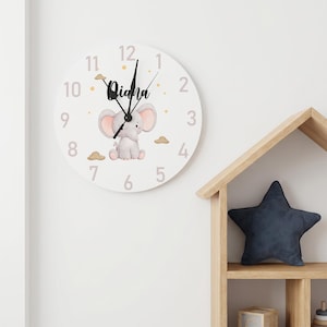 Può includere: Un orologio da parete rotondo bianco con un'illustrazione centrale di un elefante grigio con orecchie rosa, circondato da piccole nuvole marroni e puntini gialli. Il quadrante mostra il nome Diana in corsivo nero e numeri grigi da uno a dodici.