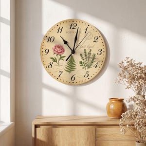 May include: A round wall clock with a cream-colored face featuring black Arabic numerals, black hands, a pink peony illustration, a green fern, and a sprig of herbs. The background includes faint text reading POSTKARTE and Dépôt légal.