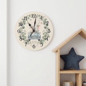 Può includere: Un orologio da parete rotondo con sfondo color crema che presenta un'illustrazione centrale di un elefante grigio alla guida di un'auto blu. Il quadrante è circondato da una corona di foglie di eucalipto verdi, piccole stelle e numeri neri. Il nome ELTO è stampato.