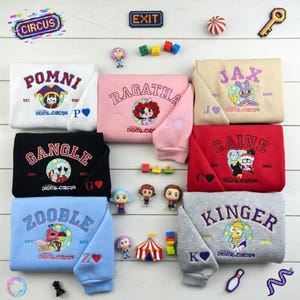 May include: A collection of folded sweatshirts in various colors, each embroidered with a character name and "Digital Circus" logo. Additional circus-themed accessories and figurines are arranged around the sweatshirts.