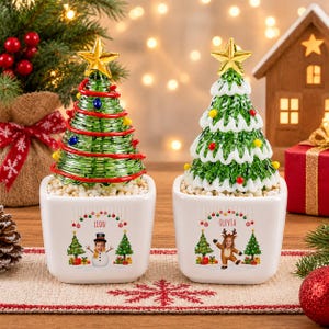 Puede incluir: Dos macetas cuadradas de cerámica blanca con gráficos navideños personalizados. La maceta izquierda muestra un muñeco de nieve y el nombre LEON, mientras que la derecha muestra un reno y el nombre OLIVIA. Cada una contiene un árbol de Navidad verde con una estrella dorada.
