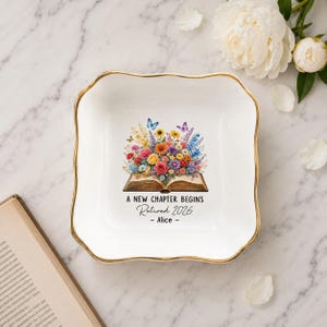 May include: A white ceramic trinket dish with a gold rim. The dish features a floral design with butterflies and the text "A NEW CHAPTER BEGINS Retired 2026 - Alice -."