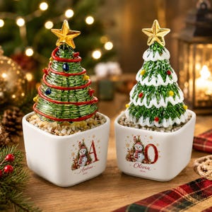 May include: Two glass Christmas tree figurines in white square ceramic pots. The left tree has red spiral garland and a gold star. The right tree has white snow-like trim and a gold star. Pots feature snowman graphics and names Andrew and Olivia.