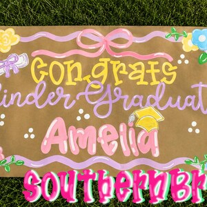 May include: A hand-painted graduation sign on brown paper, featuring the words "Congrats Kinder Graduate Amelia" in yellow and pink lettering. The sign is decorated with floral accents and a graduation cap illustration. The sign also includes the text "Southern Brush".