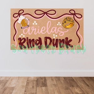 Può includere: Un cartello marrone con le parole "arielas Ring Dunk" in lettere rosa e bordeaux. Il cartello presenta illustrazioni di un boccale di birra e un anello d'oro. Sono presenti anche nastri rosa e bordeaux e testo verde.