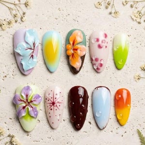 May include: Aura Botanical Collection nail art set. Ten oval-shaped artificial nails with various floral and abstract designs. Colors include lavender, yellow, orange, pink, green, and brown. Some nails feature 3D floral embellishments and water droplet details.