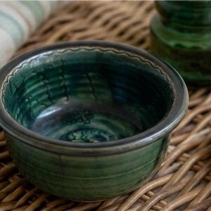 May include: A handmade, small, round ceramic bowl with a dark green glaze and a dark rim. The bowl has a textured interior and sits on a woven surface. A second green glass object is in the background.