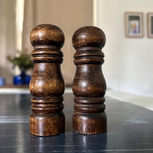 May include: A pair of dark brown wooden salt and pepper grinders featuring a turned, cylindrical design with a rounded top, a bulbous middle section, and horizontal grooved detailing along the body, showcasing a visible natural wood grain.
