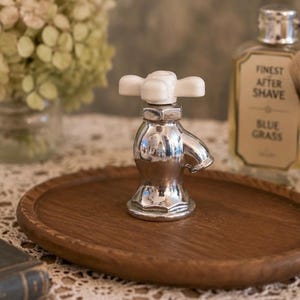 May include: A chrome-finished faucet-shaped bottle stopper with a white handle, resting on a round wooden tray. The stopper is next to a bottle of aftershave and a shaving brush, suggesting a grooming theme.