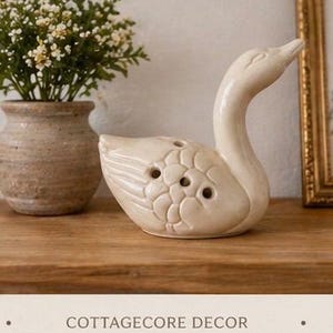 May include: A cream-colored ceramic swan figurine with a long neck and detailed feather design. The swan has several holes on its back. The text "COTTAGECORE DECOR" is at the bottom of the image. A small vase with flowers is to the left.