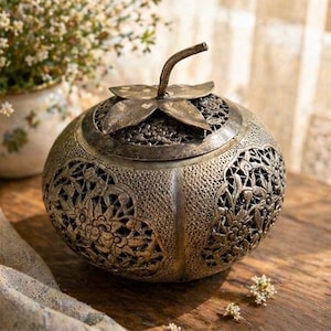 May include: A decorative, antique-style metal container with intricate floral cutouts. The container has a lid with a leaf-shaped handle. The container is silver-colored and sits on a wooden surface. A small vase with flowers is in the background.