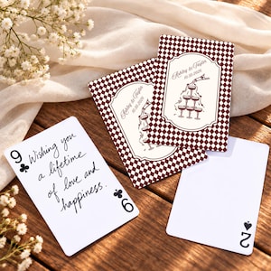 Peut inclure: Cartes à jouer personnalisées avec un motif en losanges bordeaux et blanc, une illustration de tour de champagne, le texte Ashley & Tayler 10.30.2026, et une carte avec le texte Wishing you a lifetime of love and happiness et des symboles.