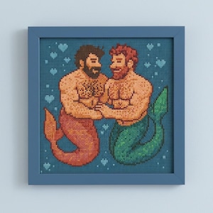 Könnte beinhalten: Kreuzstich-Kunstwerk in einem blauen Rahmen. Das Bild zeigt zwei muskulöse Meerjungmänner mit braunen und roten Haaren, die sich an den Händen halten. Einer hat eine rote Schwanzflosse, der andere eine grüne. Der Hintergrund ist blau mit kleinen weißen Herzen.