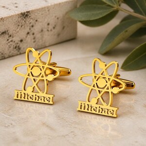 May include: A pair of gold-toned metal cufflinks featuring an atomic symbol design with three intersecting elliptical orbits and a central nucleus. Below the atom, a rectangular bar displays the name Michael in a stylized, decorative font.