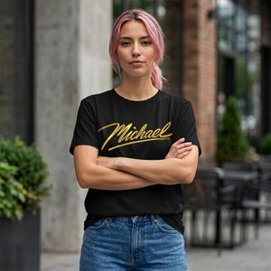 Op de afbeelding: Zwart T-shirt met de naam "Michael" in gouden letters. Het shirt wordt gecombineerd met een blauwe spijkerbroek. De persoon staat buiten met gekruiste armen. De achtergrond toont een bakstenen gebouw.