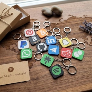 Könnte beinhalten: Eine Ansammlung quadratischer Schlüsselanhänger mit Logos von Social-Media- und Technologieunternehmen, darunter WhatsApp und GitHub. Die Schlüsselanhänger liegen auf einer Holzoberfläche mit Weltkartenmuster, neben einer Geschenkbox und einer Karte mit der Aufschrift "Unique Tech Gifts - Handcrafted."