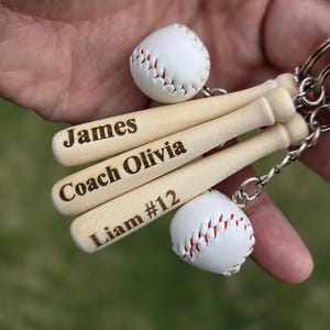 Könnte beinhalten: Baseball-Schlüsselanhänger mit Miniatur-Baseballschlägern aus Holz, graviert mit Namen und Nummern, gepaart mit kleinen weißen Basebällen mit roten Nähten. Die Schlüsselanhänger sind an silberfarbenen Ringen befestigt.