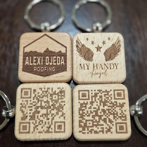 Op de afbeelding: Vier houten sleutelhangers met zilveren sleutelringen. Twee sleutelhangers hebben gegraveerde tekst: "ALEXI OJEDA ROOFING" en "MY HANDY Angel". De andere twee sleutelhangers hebben QR-codes. De sleutelhangers zijn vierkant en gemaakt van lichtgekleurd hout.